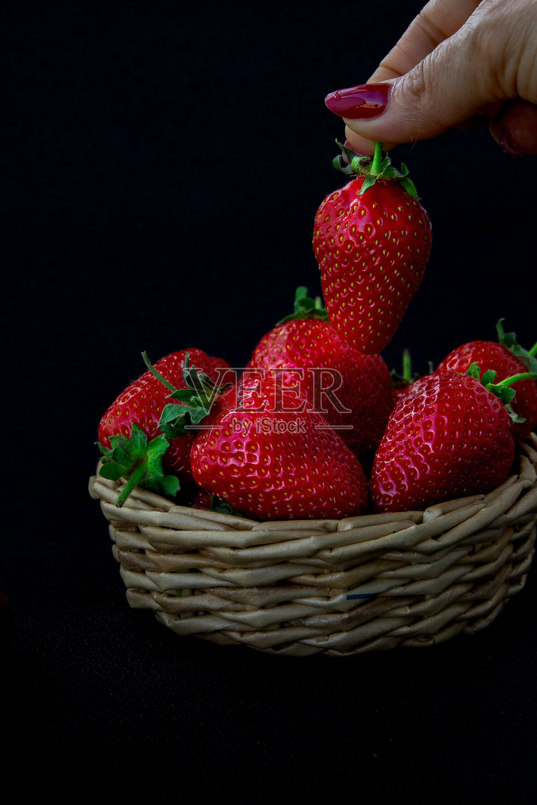 一个手工编织的篮子里装着新鲜采摘的草莓，一只手正拿起其中一颗，在深色背景下展现出鲜艳的红色。照片摄影图片