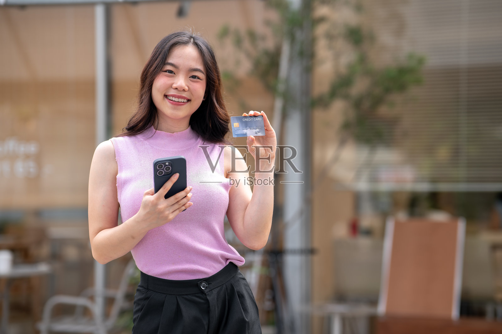 年轻的亚洲女性站在咖啡馆前或边走边看手机，并出示信用卡照片摄影图片