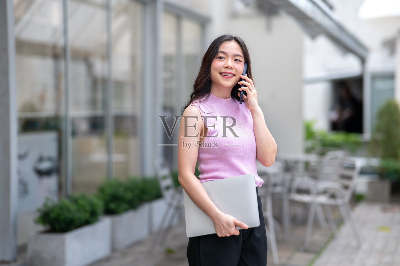 一位年轻亚洲女性一边走或一边站着，在一家咖啡馆前，一手拿着笔记本电脑，一手拿着手机正在通话。照片摄影图片