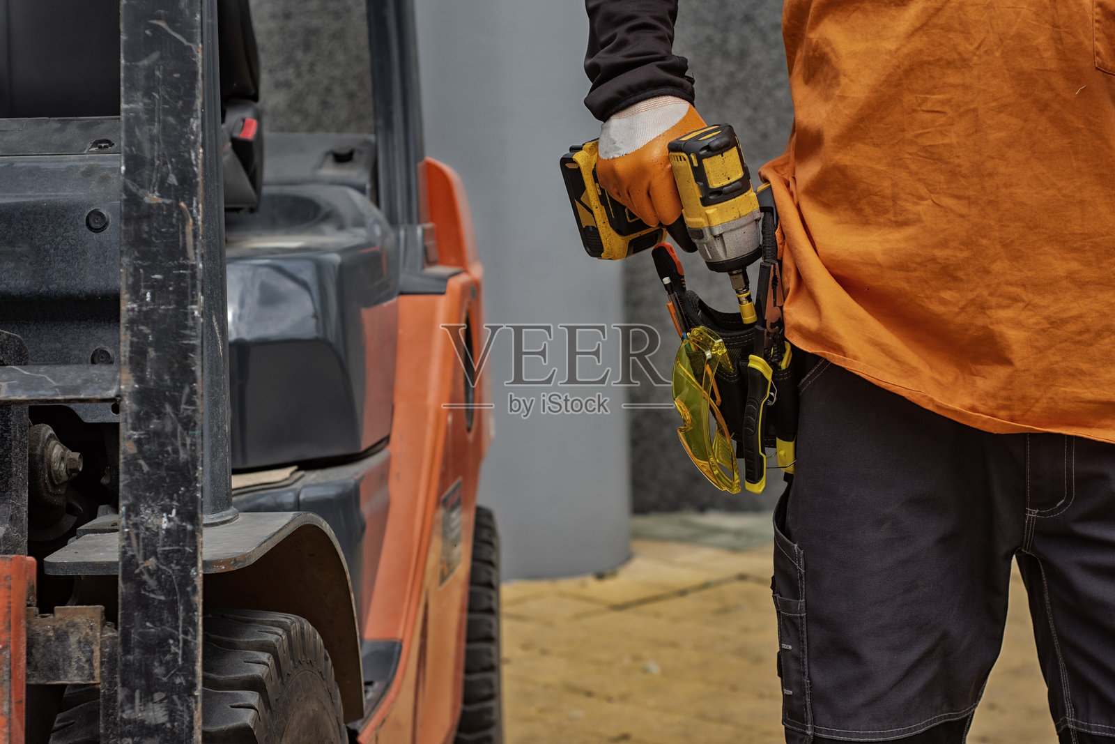 建筑工地上的一名工人，手持无绳电钻，腰间系着工具带，背景中有叉车和建筑工具。概念性专业工作，适用于工业和DIY相关内容。照片摄影图片