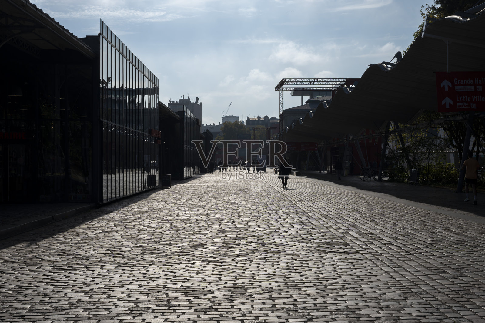 法国巴黎 - 2025年11月10日：拉维莱特公园。人们在逆光中沿着拉维莱特大厅建筑漫步在鹅卵石街道上照片摄影图片