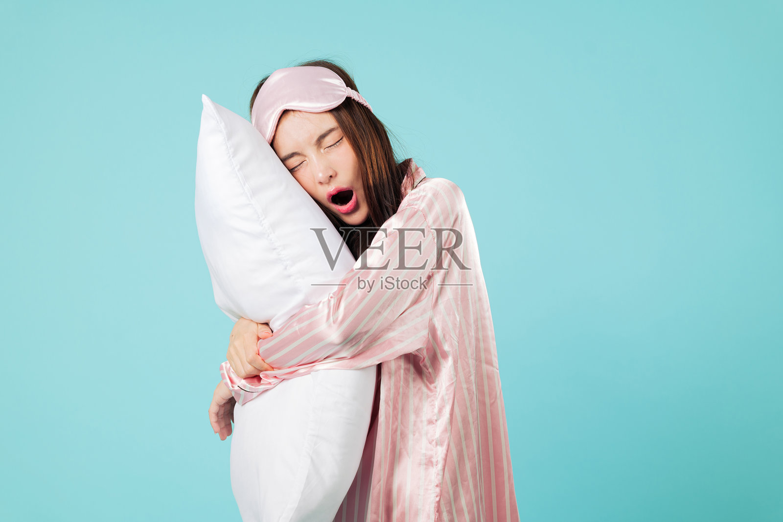 疲惫的年轻亚洲女性穿着睡衣，抱着枕头，戴着眼罩照片摄影图片