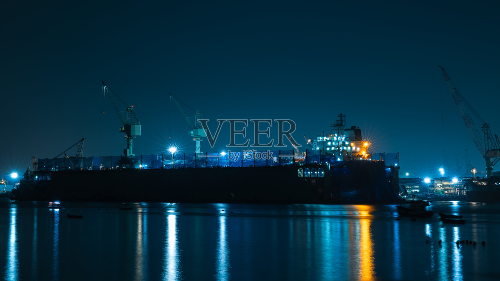 夜间大型船厂或停靠维修的船只的图像，有高耸的浅色起重机。建筑上明亮的白色和橙色灯光在深蓝色水面上投射出长长的倒影，营造出一种平静而又工业化的景象。照片摄影图片
