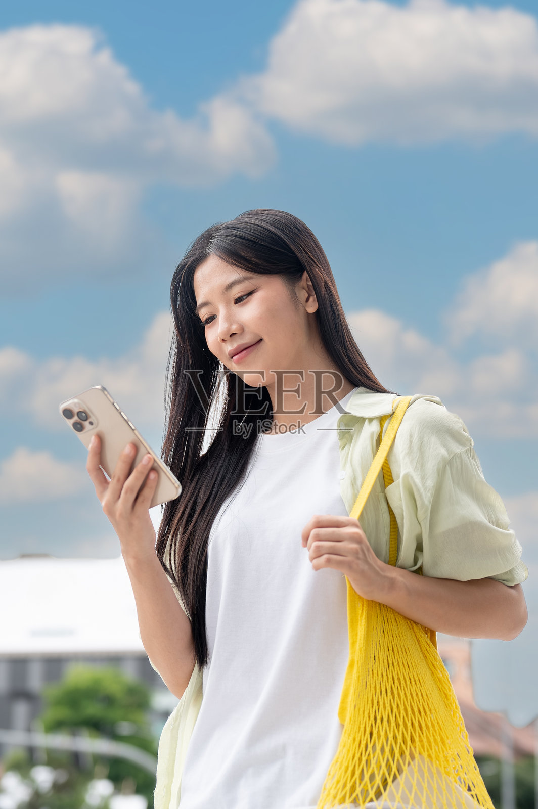 特写：亚洲女性在蓝天下户外站立或行走，看着手机，拎着包带。照片摄影图片