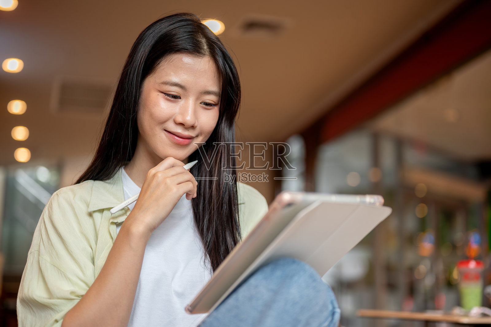 一位亚洲女学生坐在咖啡馆里，膝盖抬起，手持触控笔，看着平板电脑若有所思。照片摄影图片