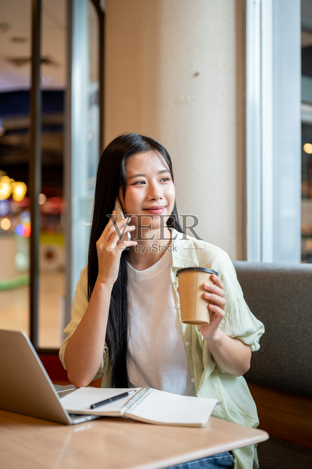 亚洲女学生在咖啡馆的木桌上，一手拿着咖啡，一手拿着笔记本和笔记本电脑，边打电话边交谈。照片摄影图片