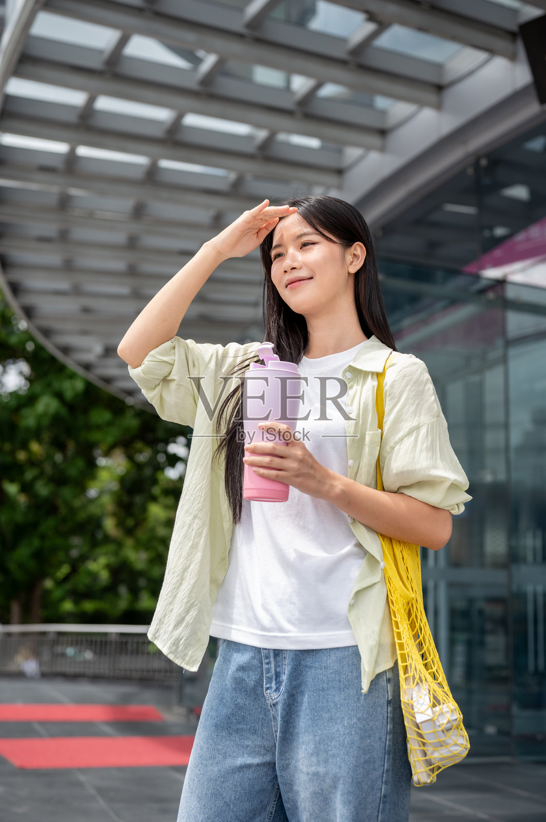 一位亚洲女性，手遮挡着阳光，手持保温瓶，站在购物中心前照片摄影图片