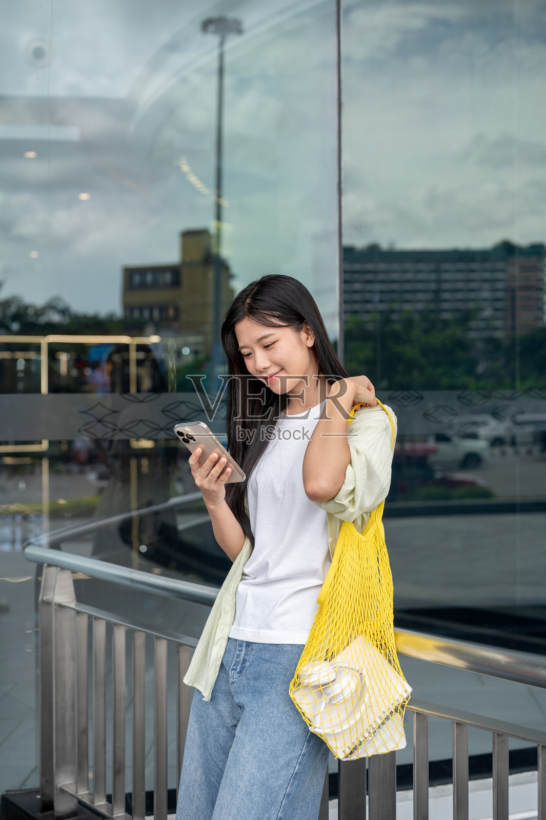 一位亚洲女性一手扶着包带，一手拿着手机，倚靠在购物商场的栏杆旁。照片摄影图片