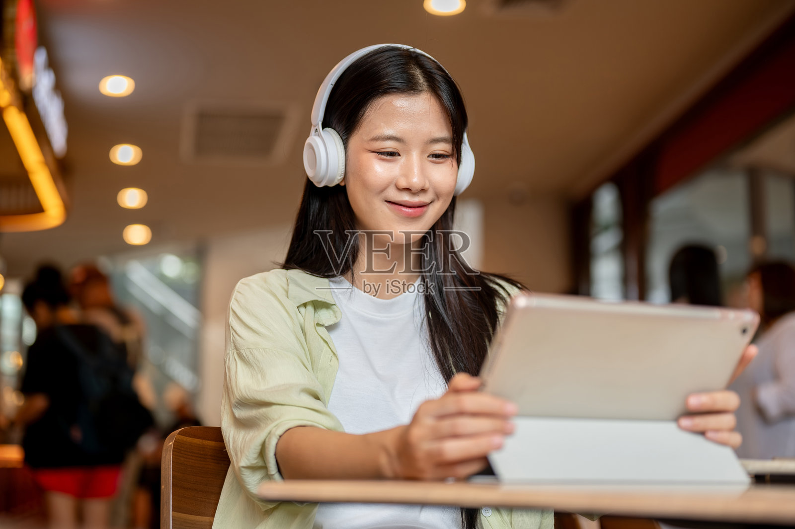 漂亮的亚洲女学生戴着耳机，在咖啡馆的木桌旁看着平板电脑。照片摄影图片