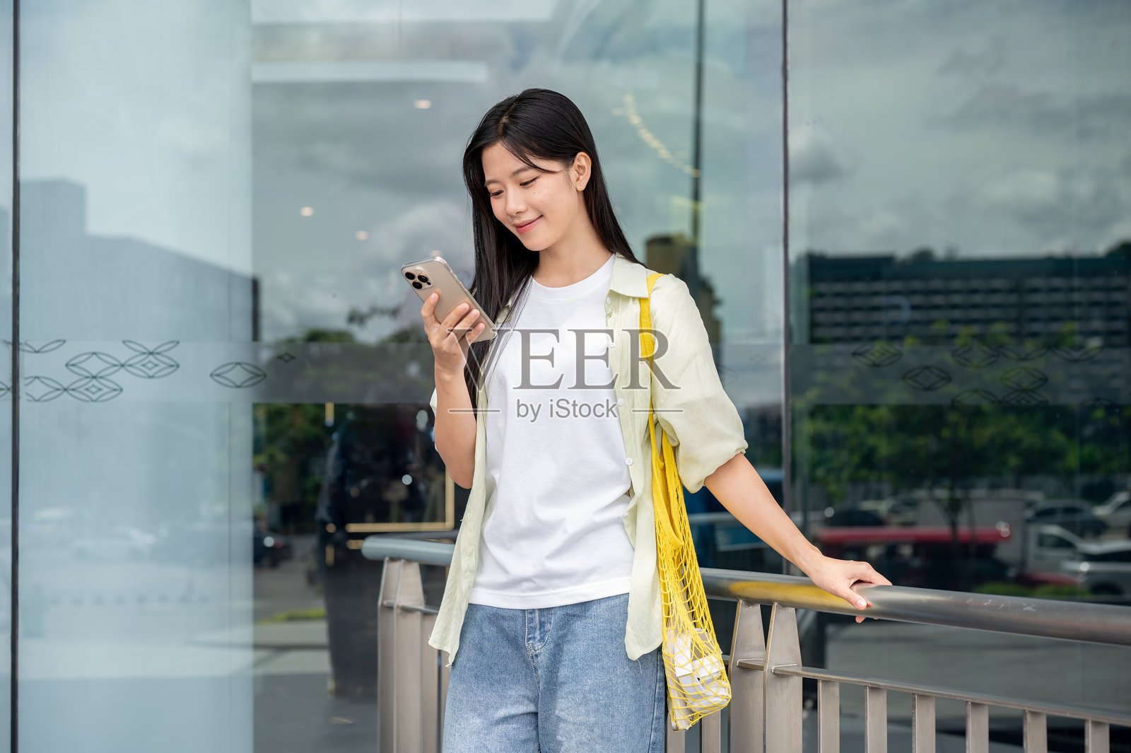 一位亚洲女性提着网袋，在购物中心的栏杆旁站着看手机。照片摄影图片