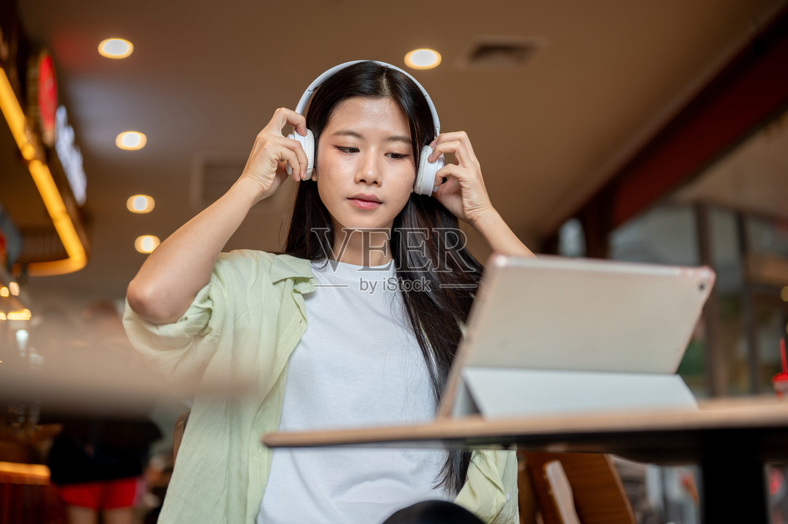 咖啡馆里，一位漂亮的亚洲女学生戴着耳机，看着桌上的平板电脑。照片摄影图片