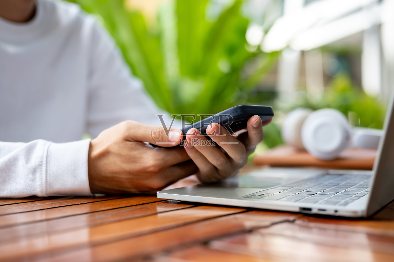 特写：男人坐在户外的木质咖啡桌旁，双手拿着手机，操作着笔记本电脑。照片摄影图片