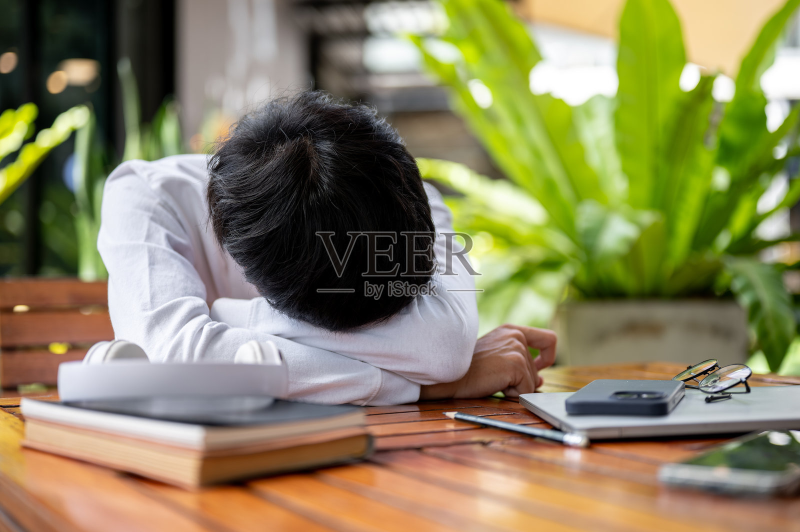 一个人在户外的咖啡馆里，趴在放有书本和手机的木桌上睡觉打盹照片摄影图片