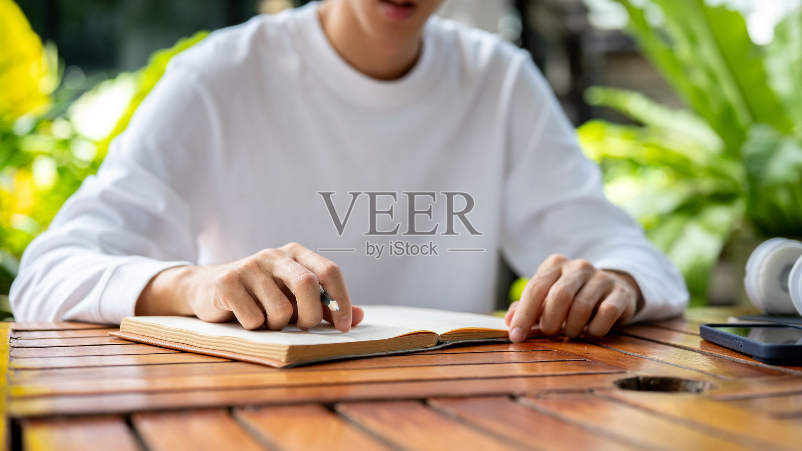 特写：男人坐在花园的木桌旁，手持铅笔，准备在笔记本上书写照片摄影图片