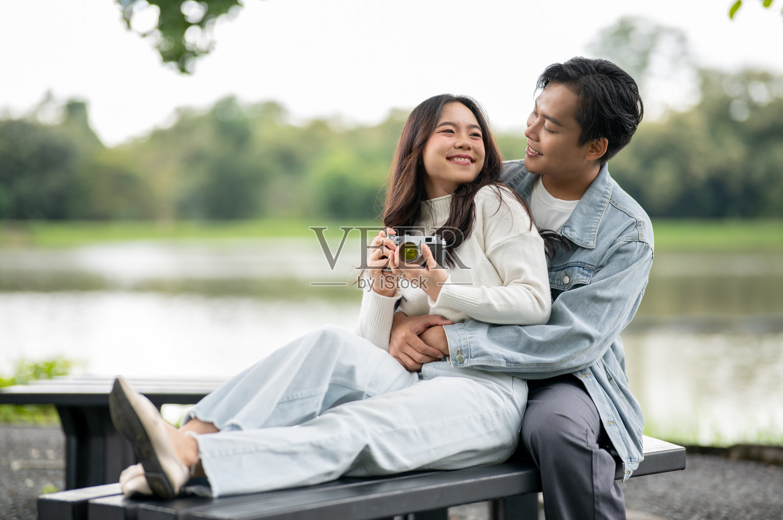 年轻的亚洲情侣，女子手持相机，男子从背后拥抱或搂着她的腰，两人坐在公园户外照片摄影图片