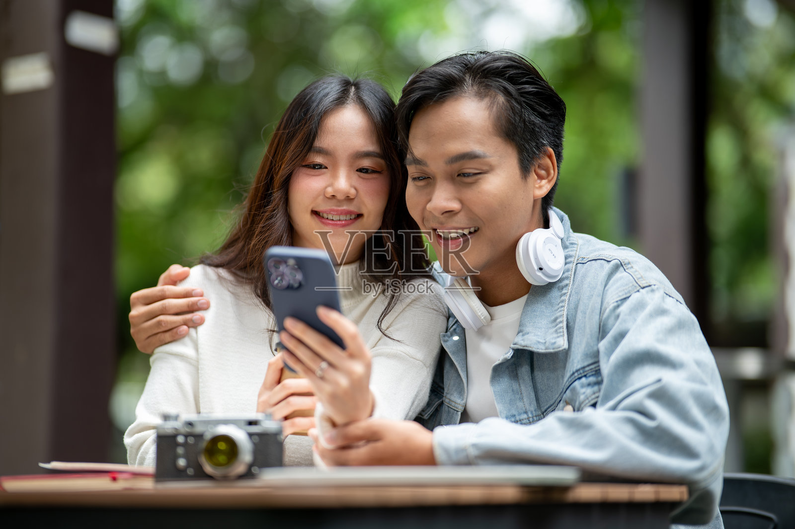 年轻的亚洲情侣，一名男子在咖啡馆的户外餐桌旁拥抱女子，同时看着手机。照片摄影图片