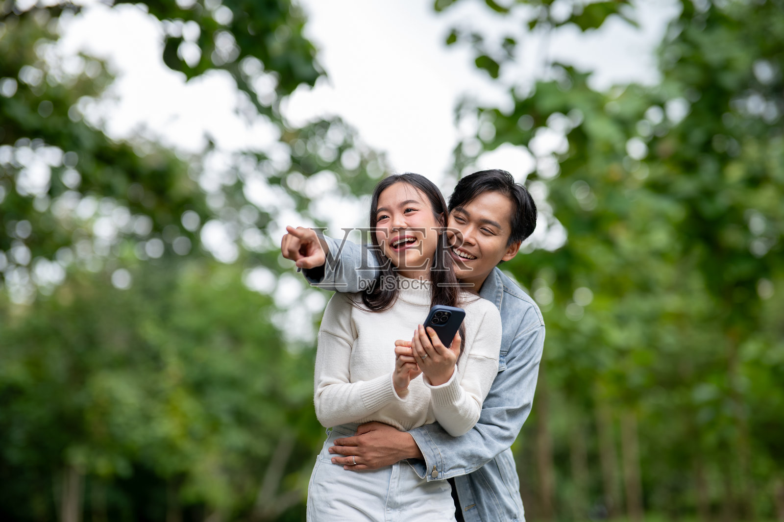 年轻的亚洲情侣，一名男子指向并拥抱一名女子，女子手持手机，两人站在公园户外。照片摄影图片