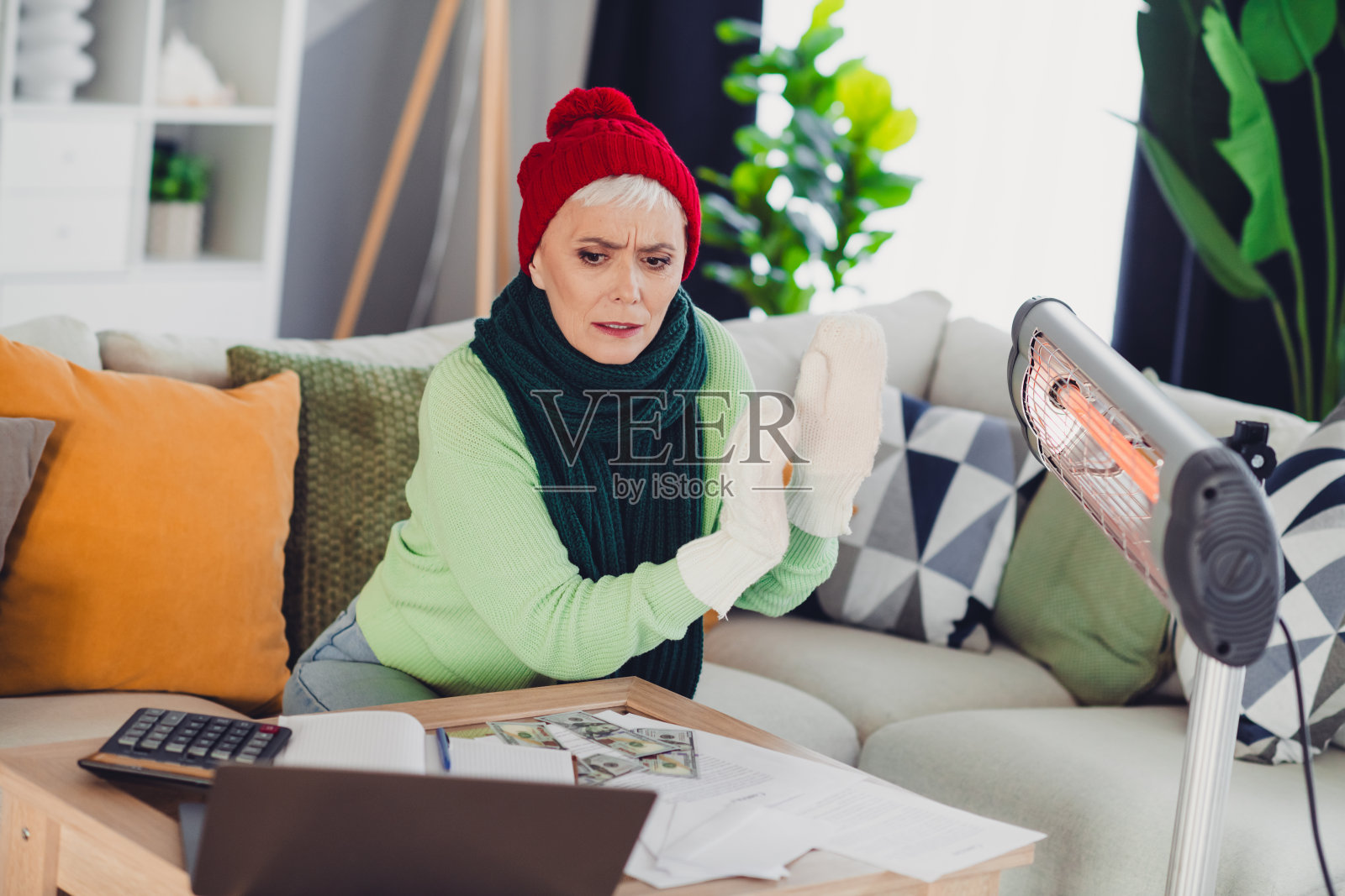 一位年长的女性在家中舒适的客厅环境里，坐在笔记本旁，用取暖器暖手，并分析账单。照片摄影图片