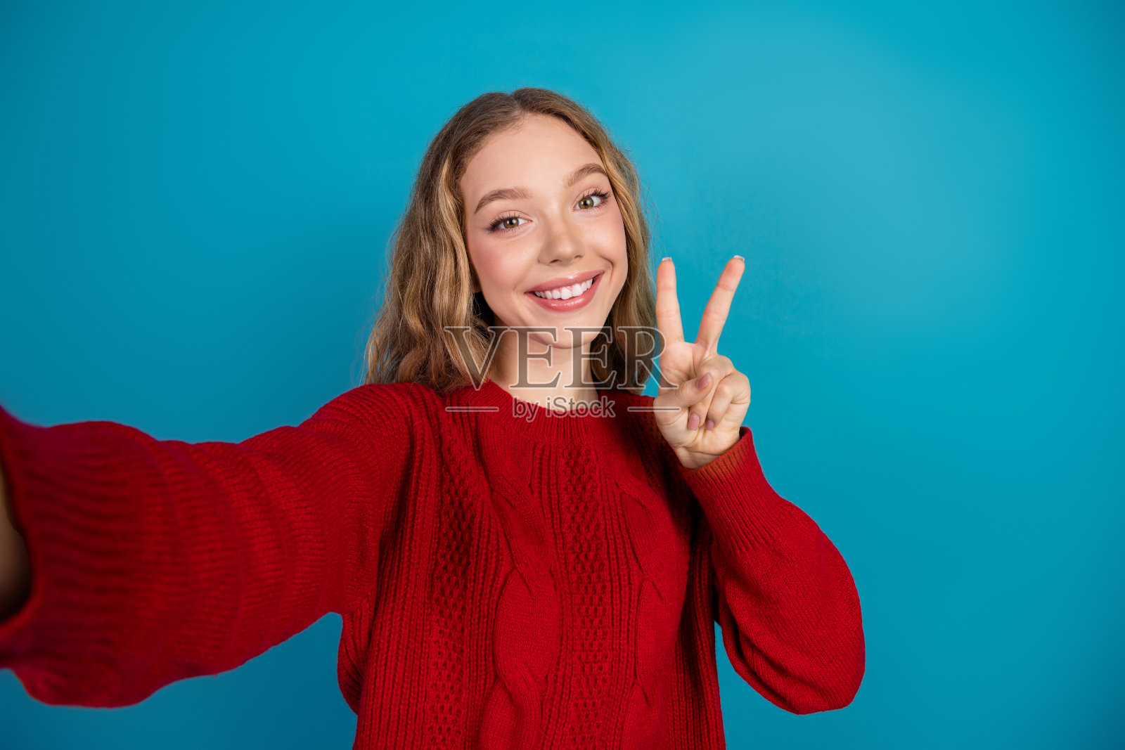一位身穿红色毛衣的年轻女子，面带微笑，在蓝色背景前摆出可爱的胜利姿势，表情愉悦。照片摄影图片