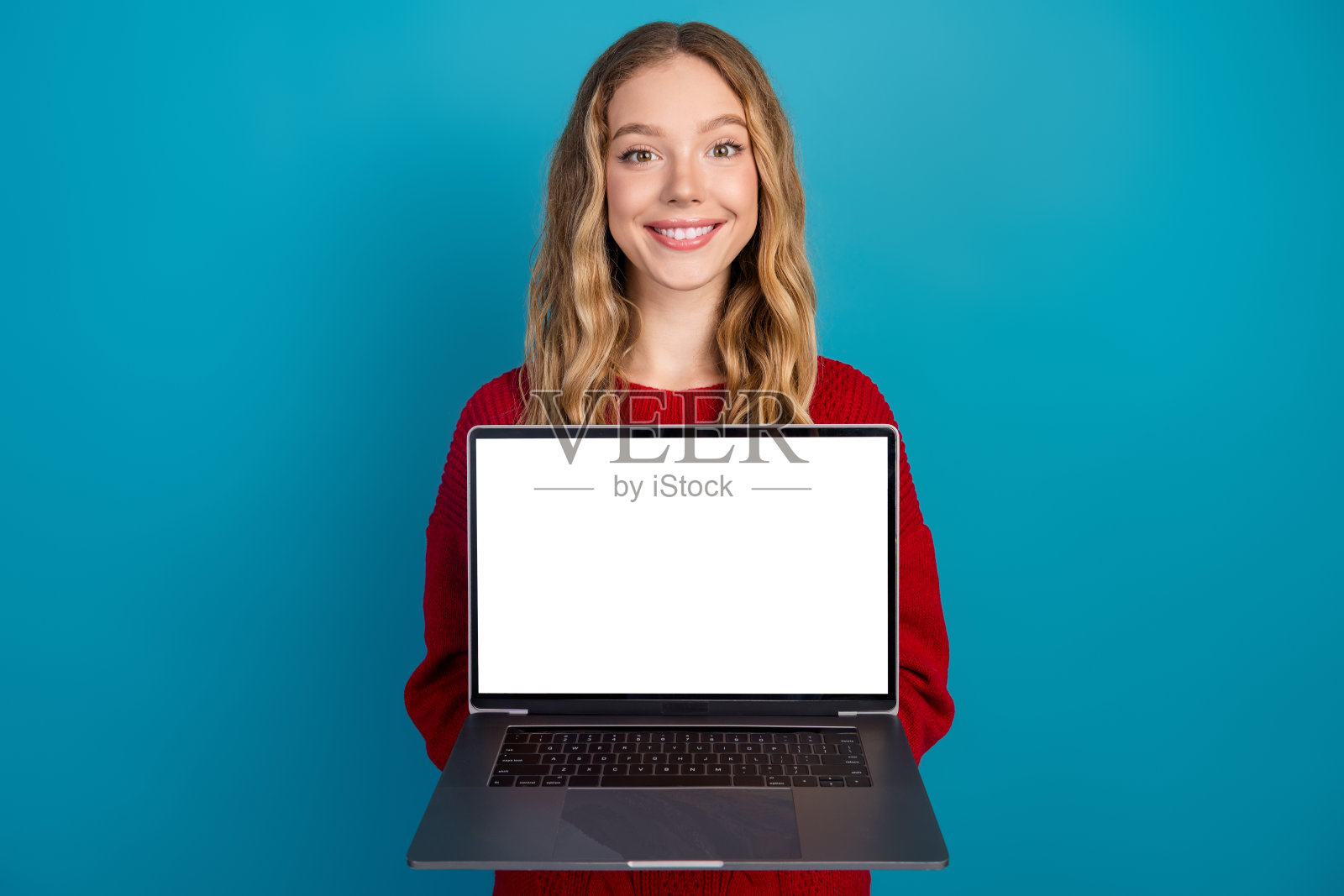 一位迷人的年轻女子穿着红色毛衣，开心地在一片蓝色背景前举着一台屏幕空白的笔记本电脑照片摄影图片