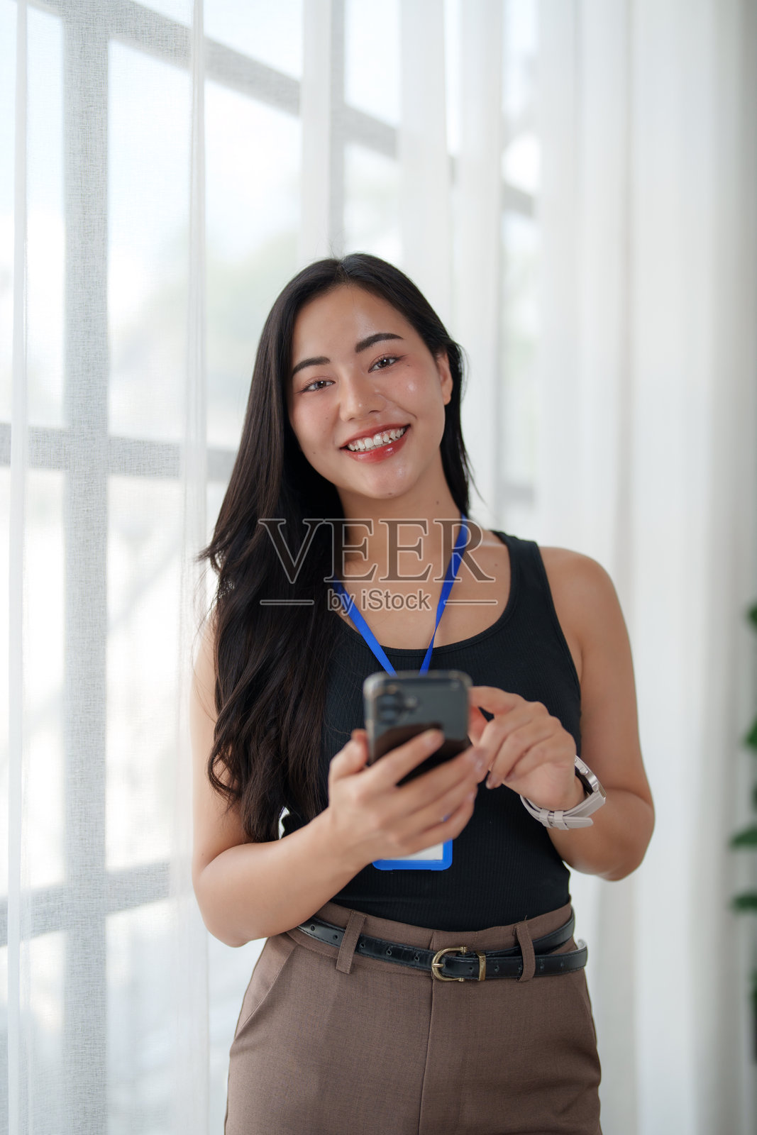 年轻的亚裔女商人对着镜头微笑，同时使用智能手机照片摄影图片