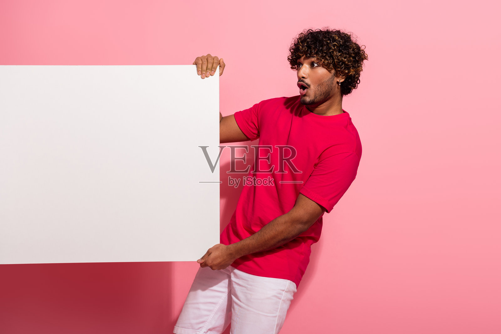 一个穿着鲜艳休闲装的年轻男子，手持空白板，在粉色背景上，脸上露出惊讶和兴奋的表情照片摄影图片