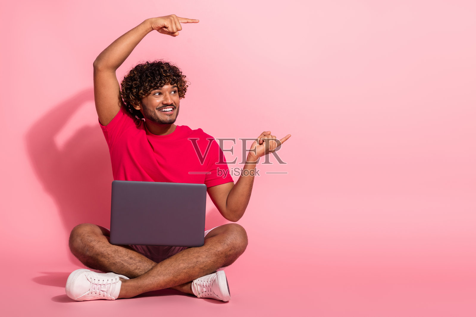 一个卷发年轻男子盘腿而坐，开心地举着笔记本电脑，指向天空，背景是充满活力的粉色。照片摄影图片