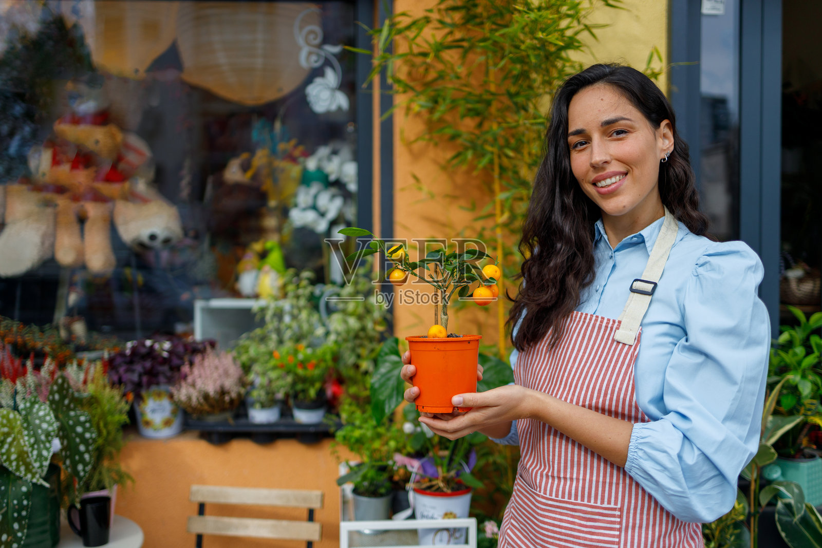 一位戴着围裙的女士微笑着，手中抱着一盆橘子树，站在她的花店前，体现了对本地小企业的支持。照片摄影图片