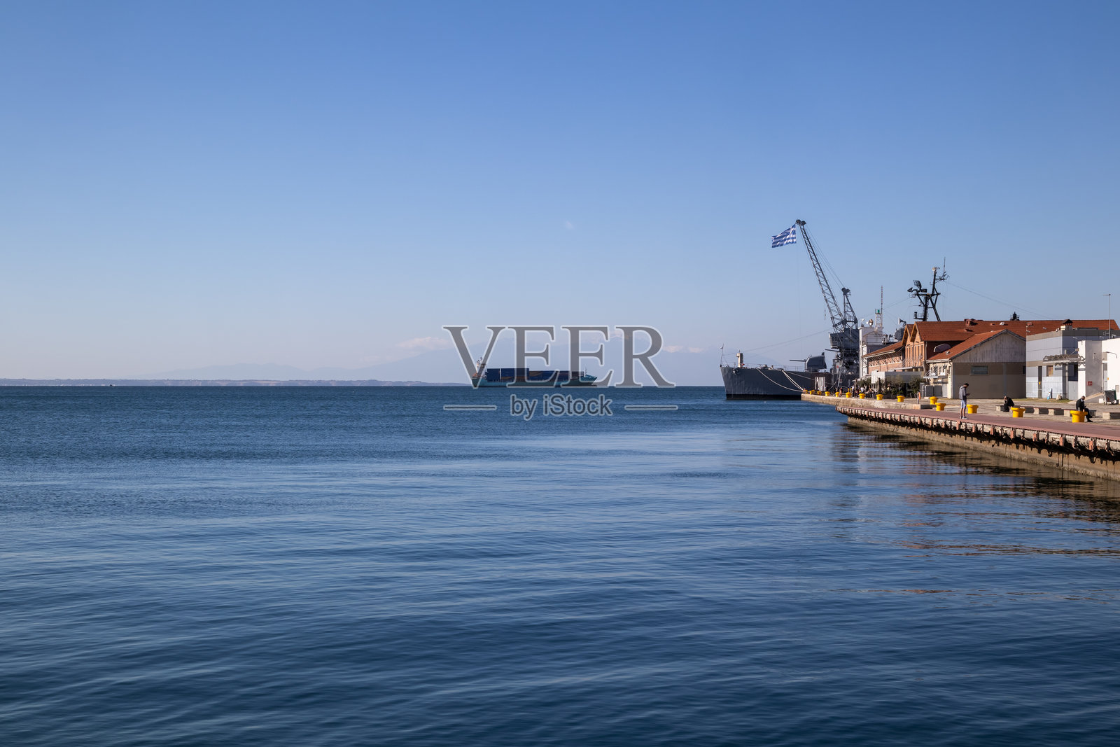 希腊港口，有集装箱船、起重机和停靠的军用或商用船只停泊在泰美科斯湾。奥林匹斯山在朦胧的远方可见，天空晴朗湛蓝。照片摄影图片