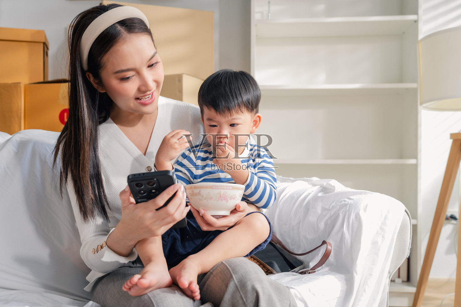 亚洲母亲一边温柔地喂儿子吃午餐，一边看着智能手机里的视频，这是他们在搬家后，从拆箱整理家具的间隙中享受的温馨家庭时光，一家人依偎在舒适的客厅沙发上。照片摄影图片