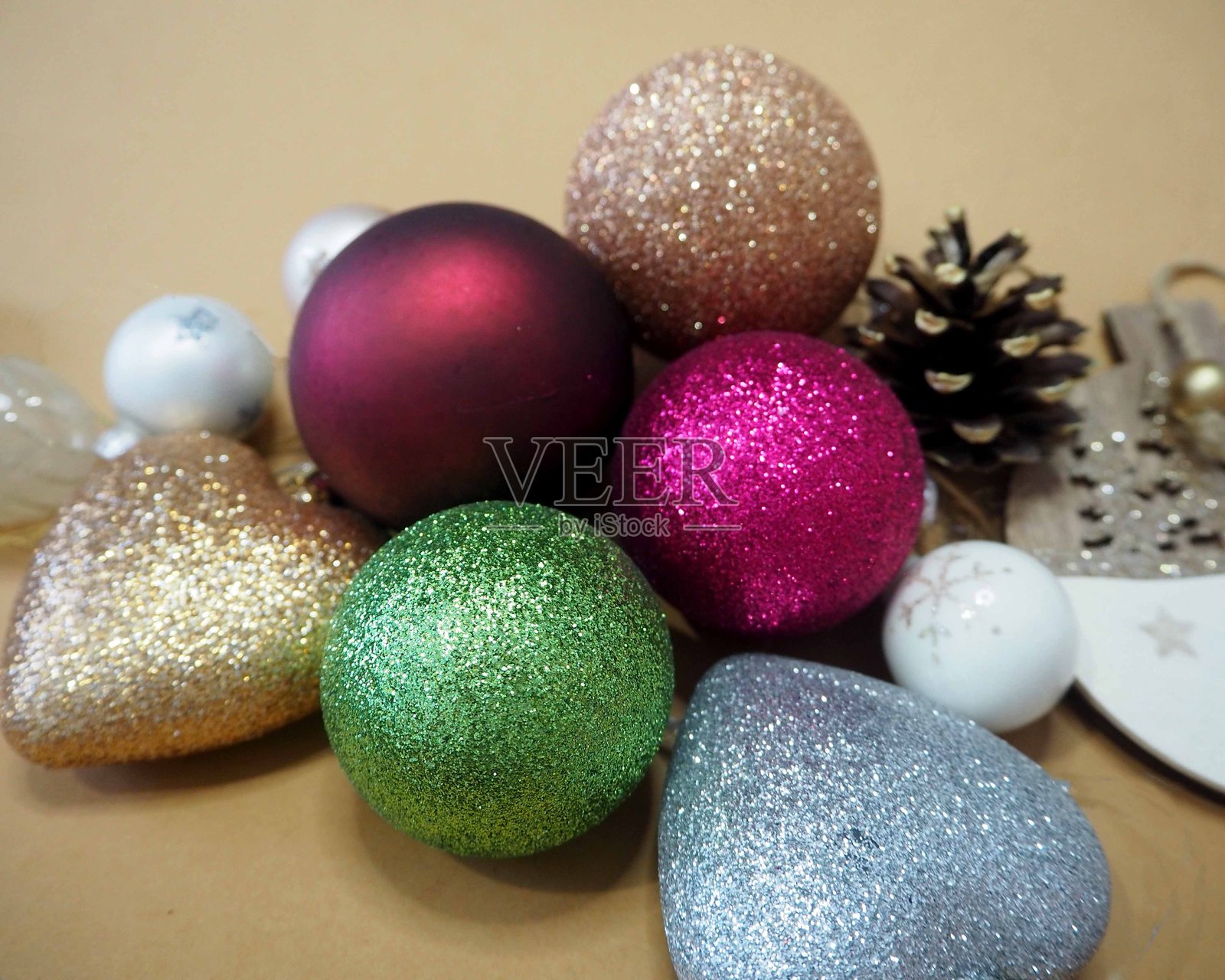 各种颜色的闪亮圣诞装饰品，包括红色、绿色、金色、蓝色和粉色，摆放在米色背景上，旁边还有松果和节日装饰。照片摄影图片