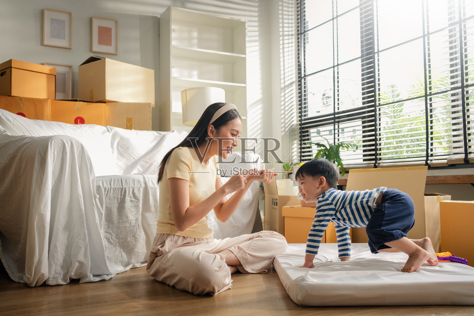 一位亚洲母亲和她活泼的儿子在搬家休息时，在地板上玩着“猜猜哪只手”的游戏，周围堆满了搬家箱，他们分享着欢声笑语、爱意和快乐的亲子时光。照片摄影图片