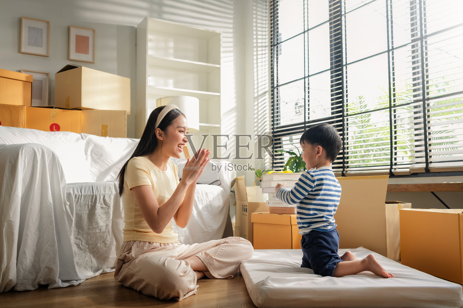 亚洲母亲和她活泼的儿子在搬家休息时，坐在客厅地板上，被搬家箱子包围，玩着“猜猜在哪只手”的游戏，分享着欢声笑语、爱意和快乐的亲子时光。照片摄影图片