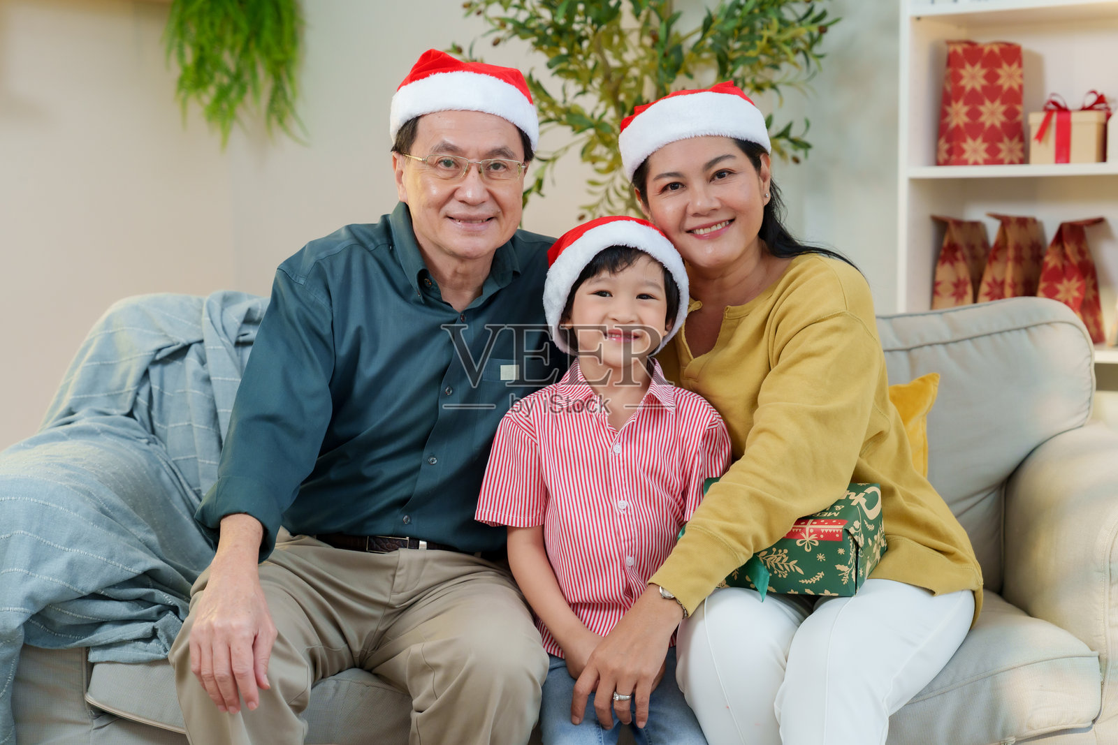 亚裔祖父母和孙子戴着圣诞帽坐在沙发上，开心地笑着，享受着圣诞节的节日家庭温馨庆祝活动，象征着幸福的节日照片摄影图片