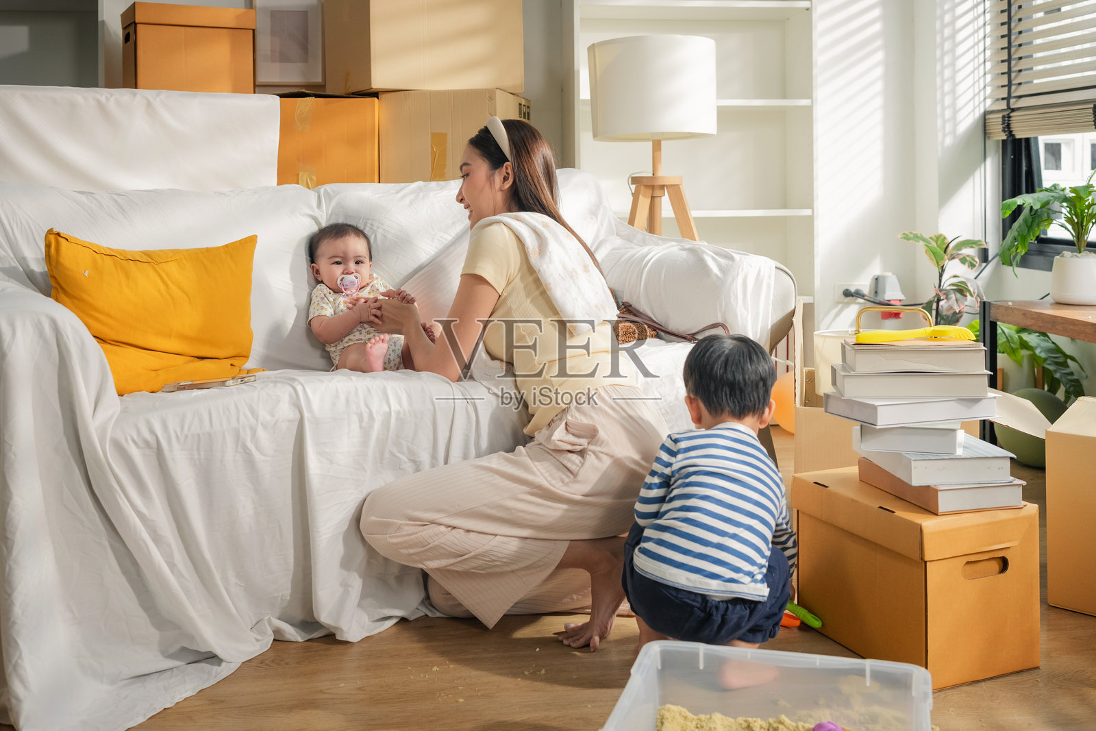 一位年轻的亚洲母亲和她的两个孩子（一个男孩和一个女婴）在搬家时，开心地拆着纸箱、布置家具，在新家安顿下来的幸福感中享受着玩耍的间歇，营造出一种宁静的生活方式。照片摄影图片