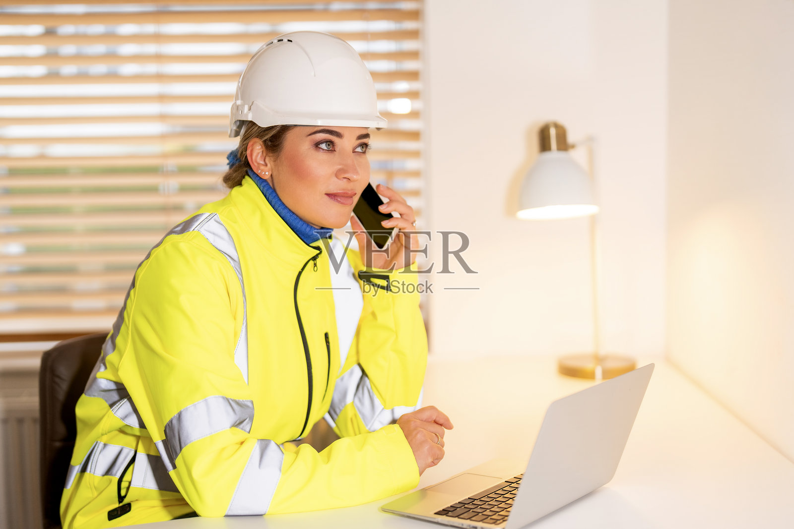 一位穿着安全外套的年轻女建筑工人，在办公室一边打电话一边使用笔记本电脑，在建筑环境中展现专业精神照片摄影图片