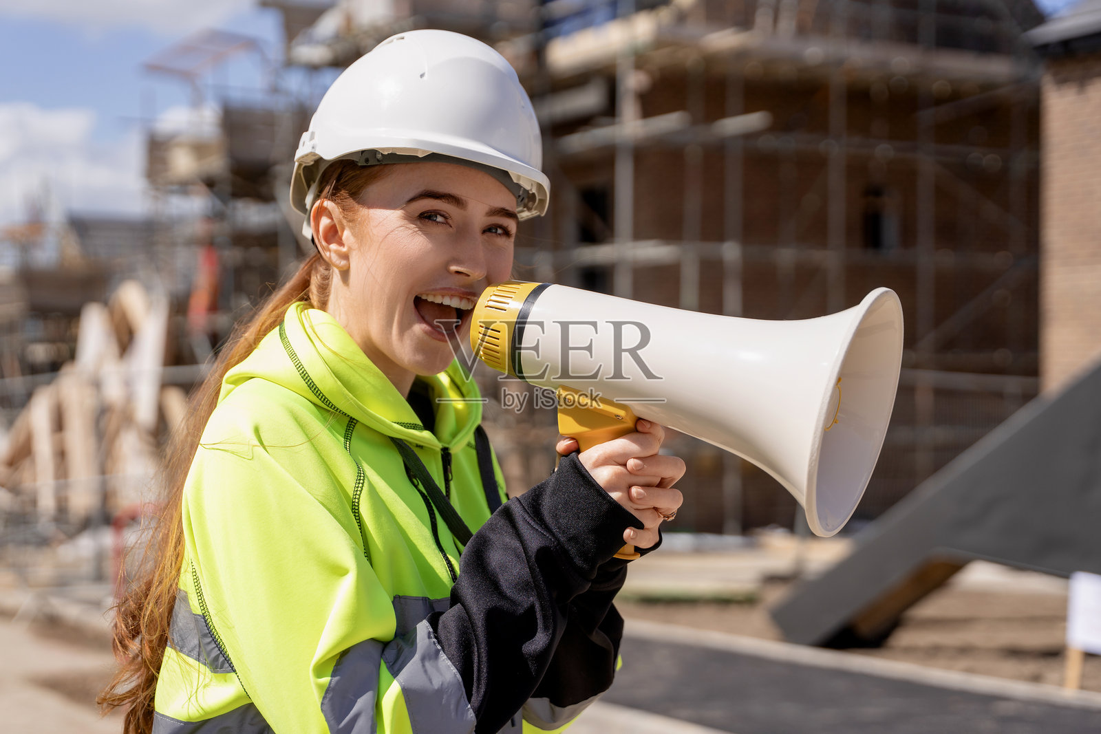 身穿橙色夹克的女建筑工人在建筑工地上用扩音器大声喊叫以吸引注意力照片摄影图片