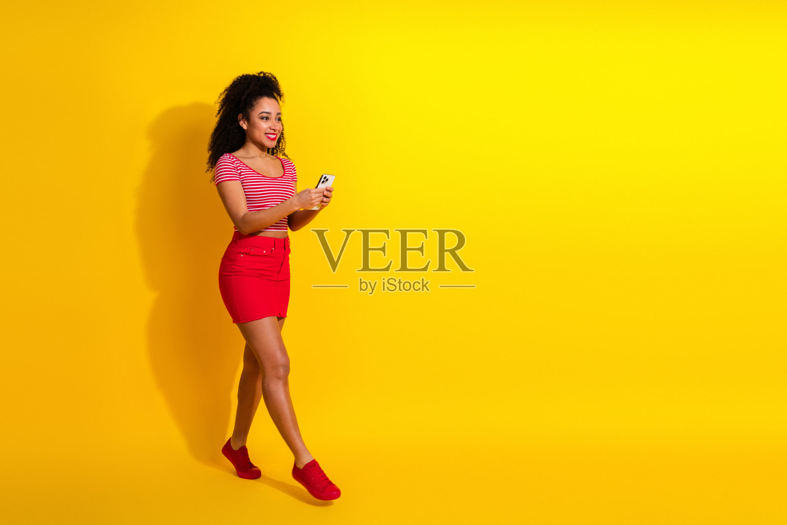 一位年轻时尚的女士，对着生机勃勃的黄色背景，喜悦地微笑着，拥抱休闲潮流，正在使用智能手机。照片摄影图片