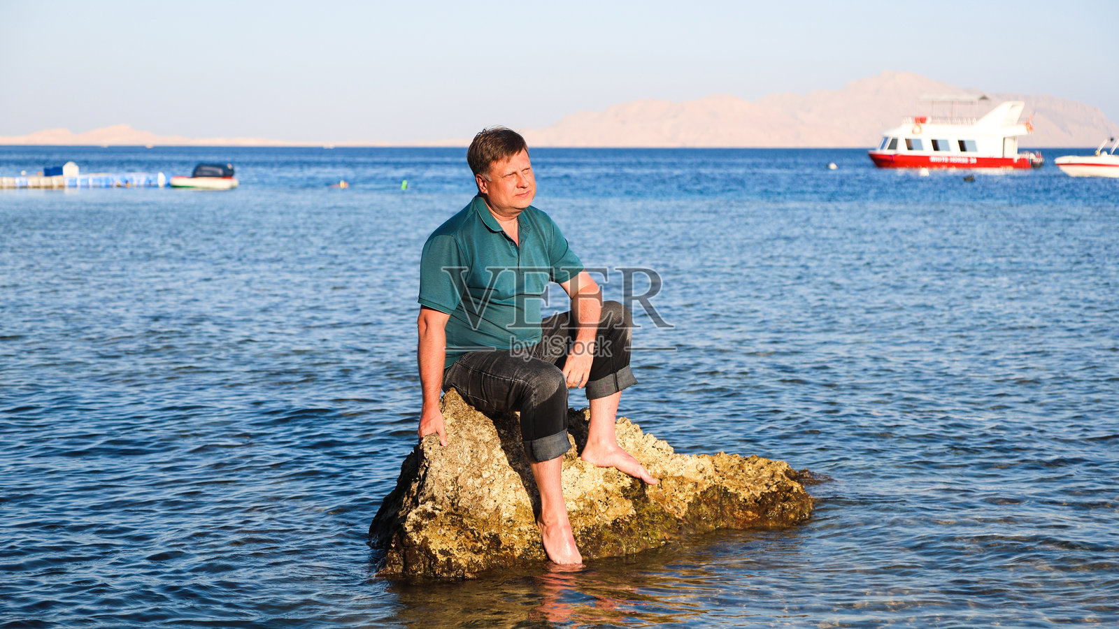 一个金发男子身着衣物，坐在海中的岩石上，远离海岸。照片摄影图片