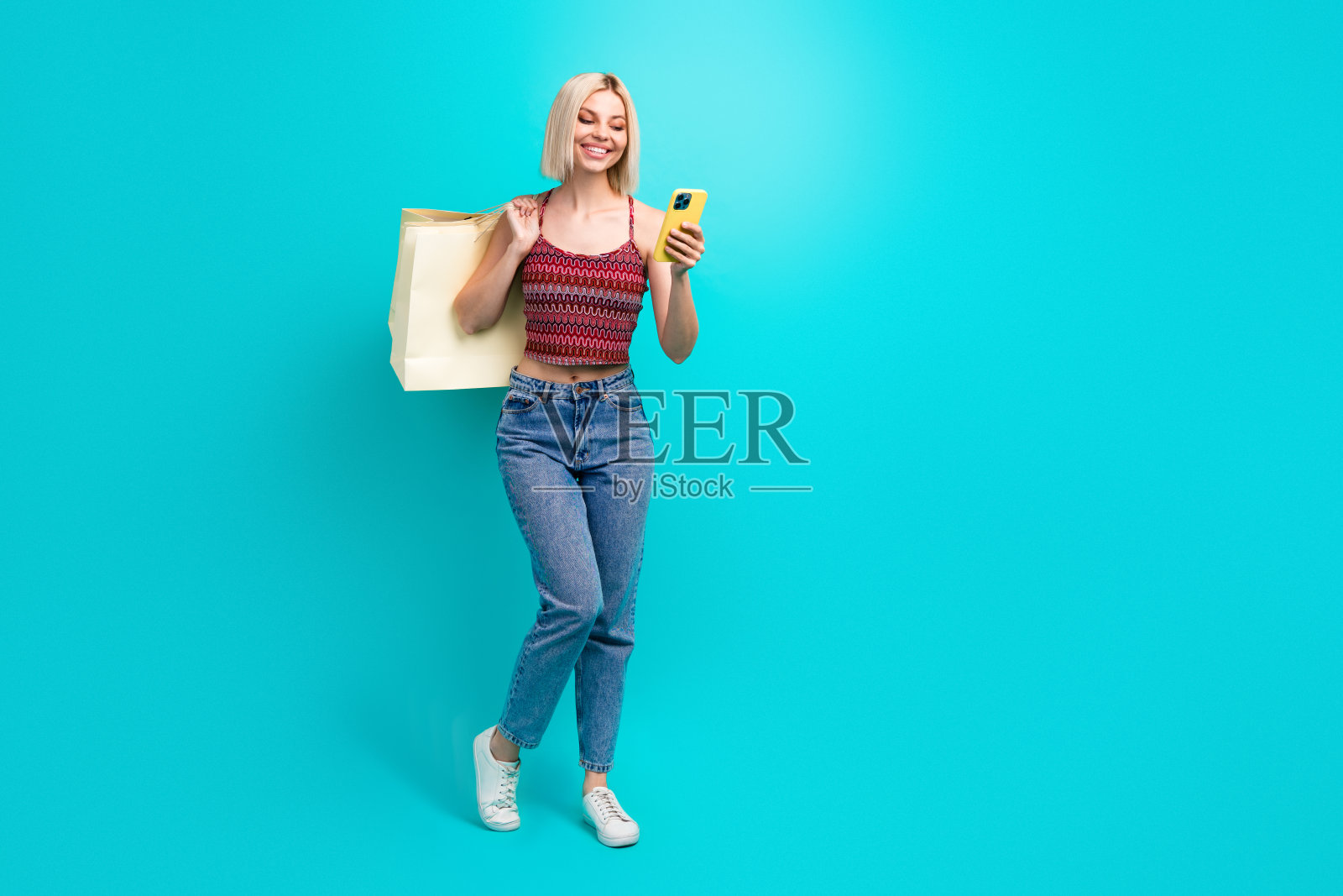 年轻女子提着购物袋，手持手机，面带微笑，身处绿松石色的背景前，展现了时尚购物和休闲风格。照片摄影图片