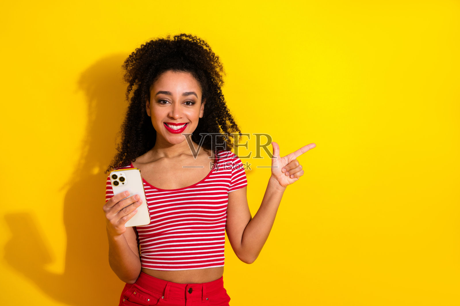 一位快乐的年轻女子手持智能手机，竖起大拇指向前指，她身处充满活力的黄色背景前，以便留白。照片摄影图片