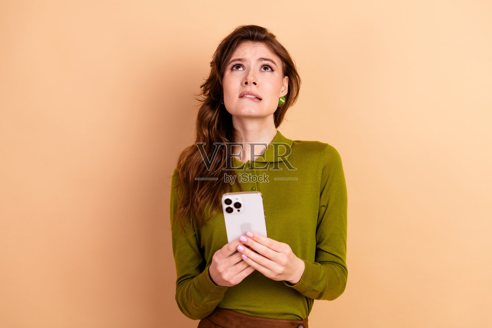 一位棕发年轻女子手持智能手机，满怀希望地抬头仰望，背景为米色，适用于生活方式时尚广告和购物概念照片摄影图片