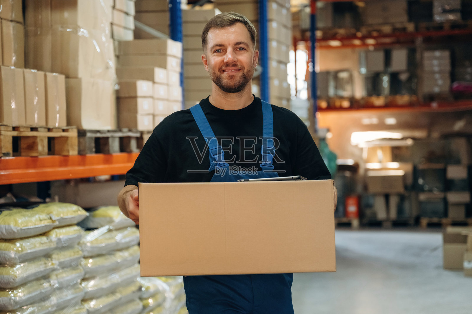 身穿蓝色制服的男子，手持箱子，正在仓库打包准备发货的包裹。照片摄影图片