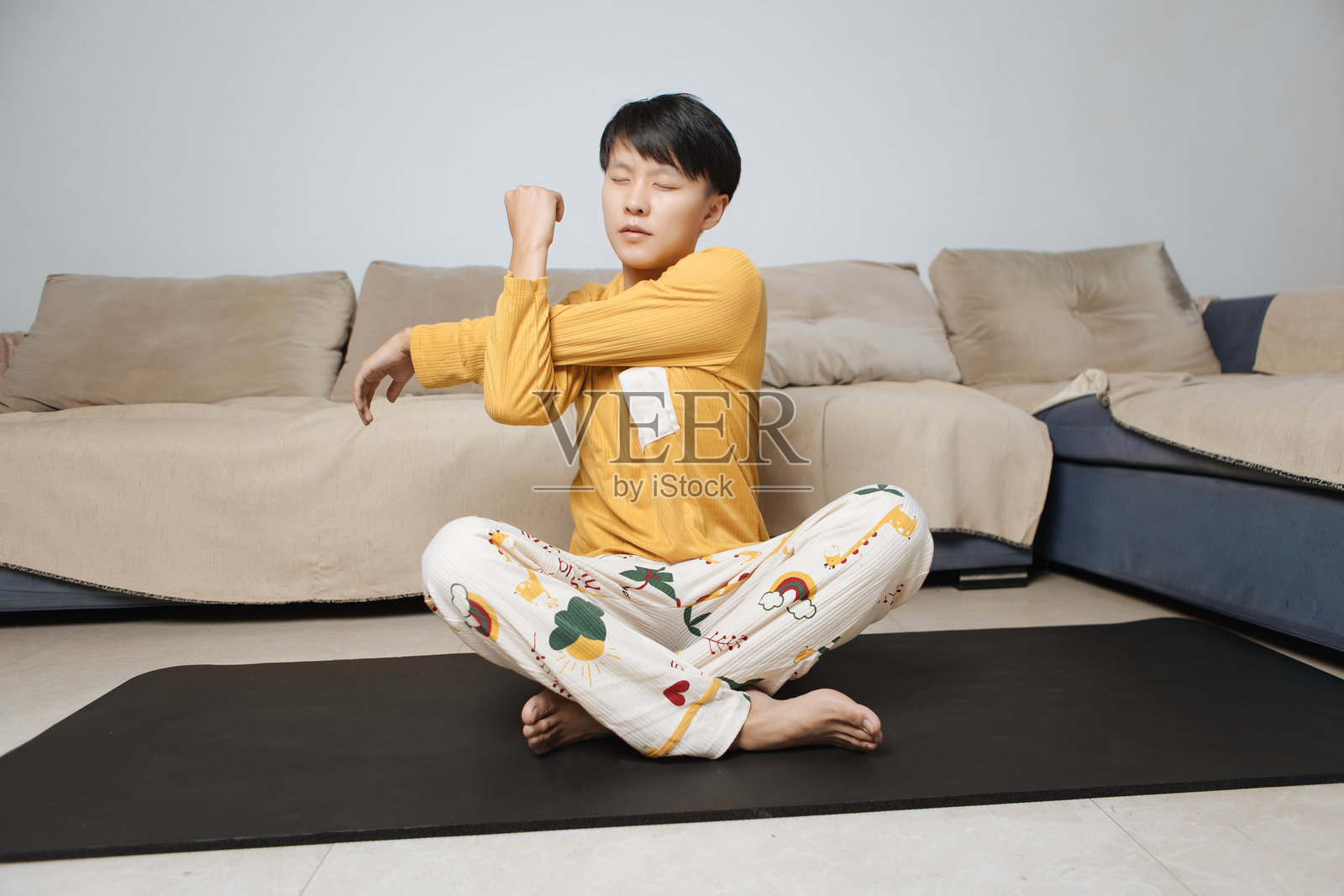 年轻的亚洲男性坐在客厅的瑜伽垫上，盘腿而坐，戴着耳机伸展肩关节，践行健康生活方式和身体护理，专注于明亮现代家居中的个人健康和室内健身。照片摄影图片