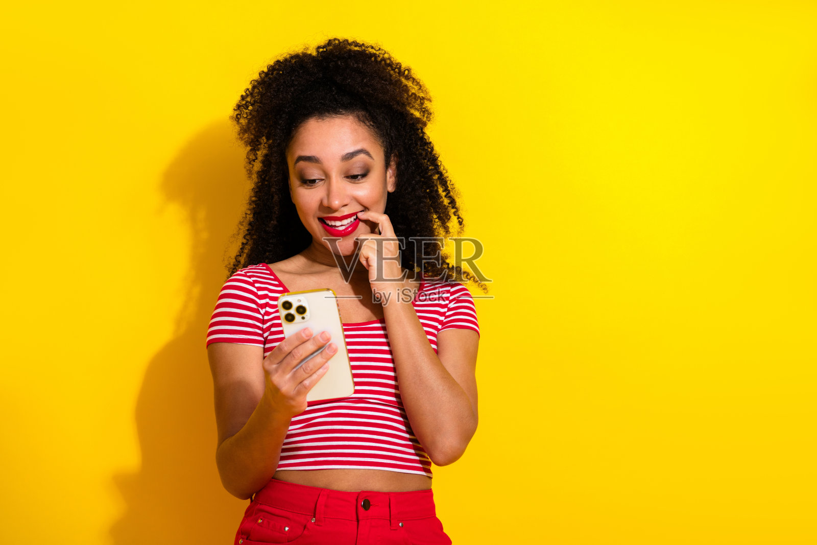 穿着条纹上衣的漂亮女子手持智能手机，背景为明亮的黄色，她带着俏皮的姿势，脸上洋溢着快乐和魅力。照片摄影图片