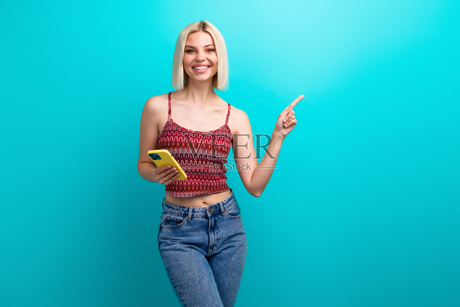 一位年轻的金发女子身穿休闲时尚的服装，站在蓝绿色背景前，手持黄色智能手机，面带微笑，同时指向一侧。照片摄影图片