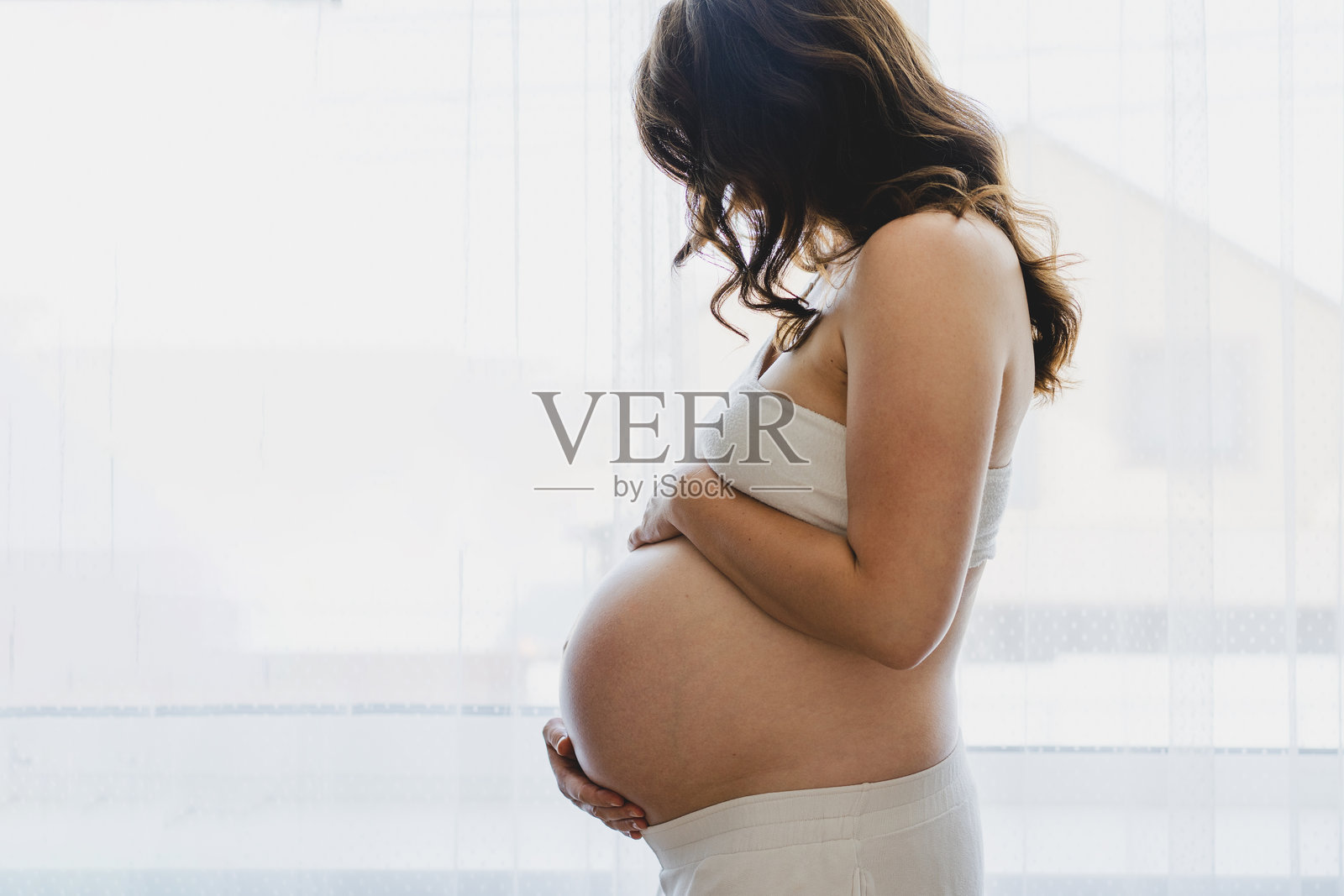 一位孕妇微笑着，在窗边抚摸着自己的肚子照片摄影图片