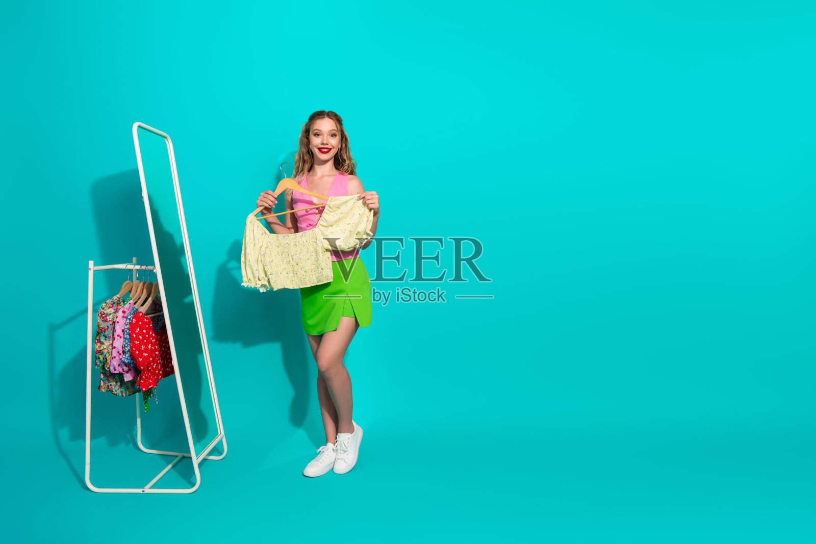 年轻时尚女子身穿粉色上衣和绿色裙子，手持黄色上衣，站在衣架旁，背景为青绿色调的影棚，非常适合购物和时尚主题。照片摄影图片