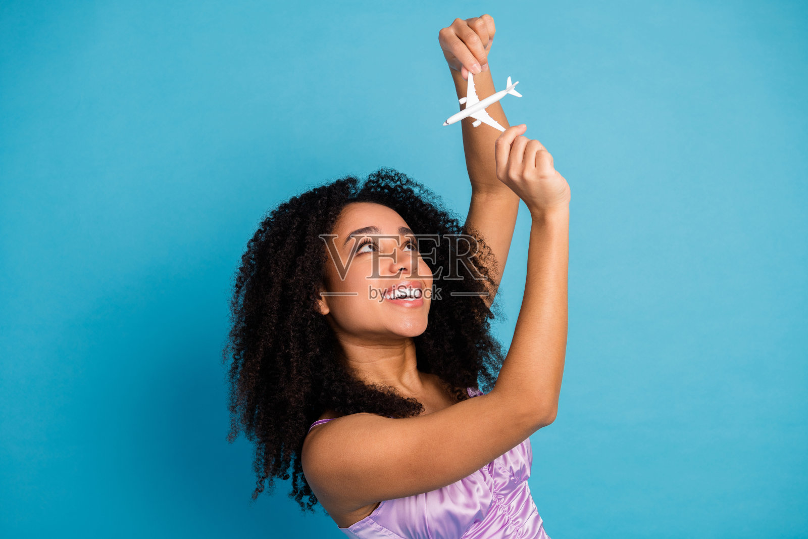 一位卷发女子高举玩具飞机，背景为蓝色，庆祝旅行梦想与俏皮时尚时刻照片摄影图片