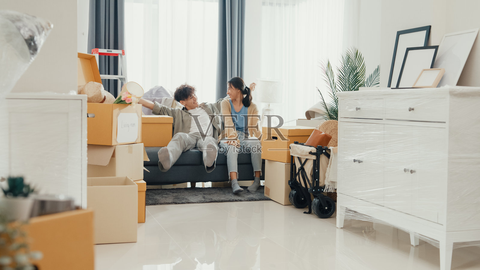 一对年轻快乐的亚洲情侣手牵手走在新房里，一起坐在客厅的沙发上。夫妻搬入新家，公寓租赁与所有权。照片摄影图片